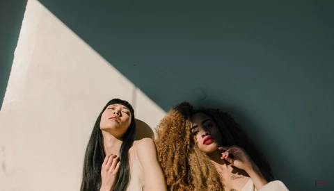 Two women with long hair looking upward against a teal wall with dramatic window light.