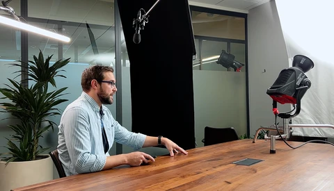Man seated at wooden desk in professional studio setup with professional lighting equipment and microphone visible.