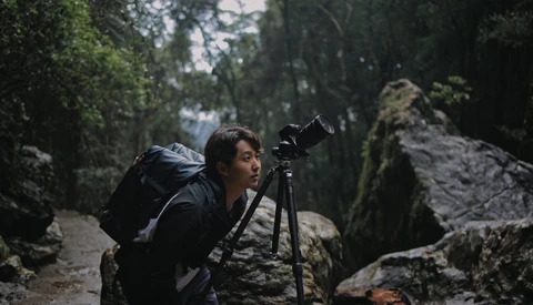 Photographer with backpack and tripod mounted camera on a forested trail surrounded by moss-covered rocks.