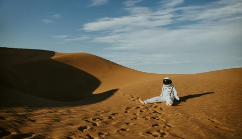 Astronaut in white spacesuit sitting alone on golden sand dune with footprints visible behind.
