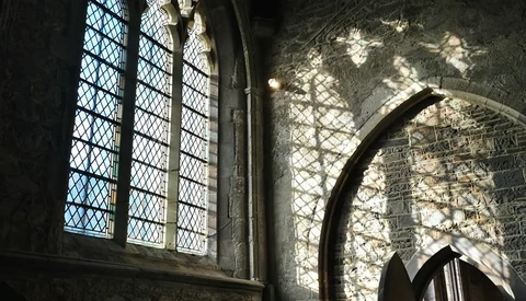 Gothic stone interior with arched doorway and latticed window casting dramatic shadows on textured walls.