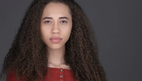 Portrait of a young woman with long curly dark hair wearing a red ribbed top against a neutral gray background.