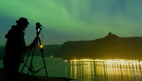 Photographer on tripod capturing northern lights over illuminated lakeside landscape at night.