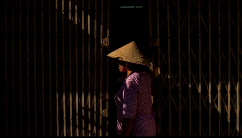 Person wearing traditional cone hat and purple garment standing in dramatically lit narrow corridor with wooden walls.