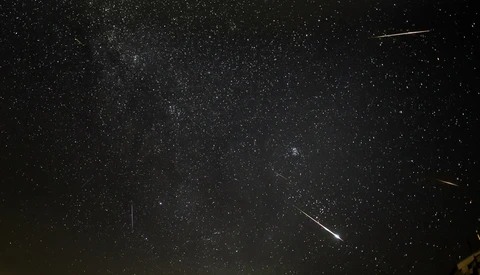 Milky Way galaxy stretching across night sky with multiple meteor streaks and silhouetted mountain ridge below.