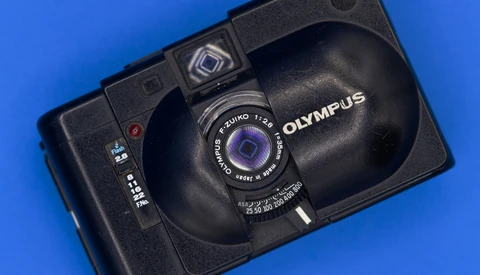 Black Olympus compact camera with Zuiko lens against blue background.