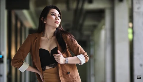 Woman in brown blazer and black top standing in modern corridor, looking upward with contemplative expression.