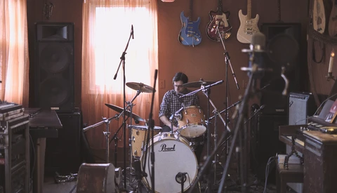 Drummer performing in a home studio surrounded by guitars and musical equipment with warm window lighting.