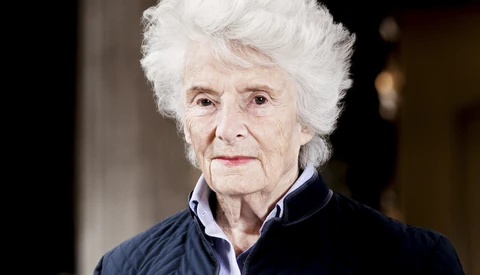 Elderly woman with white hair wearing dark jacket over light purple shirt, photographed against neutral background.