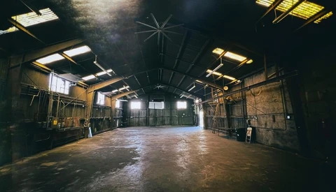 Large abandoned warehouse interior with industrial ceiling, metal scaffolding, and scattered windows casting warm light across concrete floor.