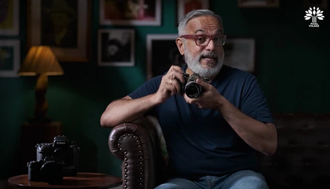 Bearded man with glasses holding a vintage camera while seated in a studio setting.