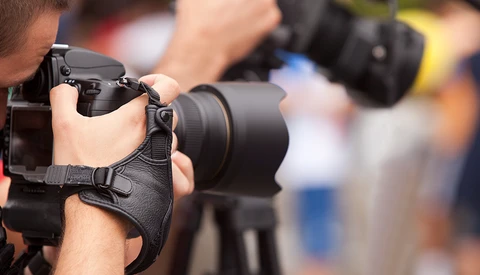 Photographer holding DSLR camera with telephoto lens and wrist strap during outdoor assignment.