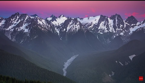 Snow-capped mountain range at dusk with a river valley and forested slopes below.