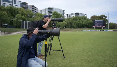 Two photographers operating large telephoto lenses on tripods at a sports field during daytime.