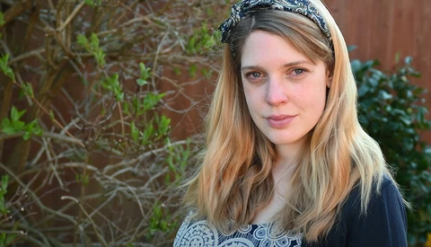 Portrait of a young woman with long blonde hair and a decorative headband against a brick and foliage background.