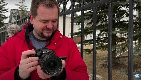 Photographer in red jacket holding a Lumix camera outdoors near a black metal fence.