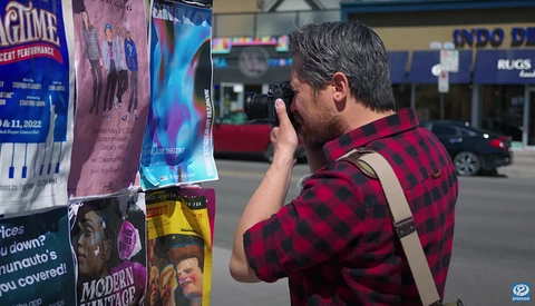 Photographer in red plaid shirt holding camera to face, composing a shot of colorful event posters on an urban street.