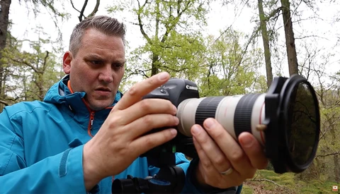Photographer in blue jacket examining camera with telephoto lens in wooded setting.