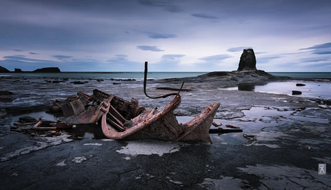 Rusted shipwreck remains on a rocky shore at twilight with a sea stack in the background.
