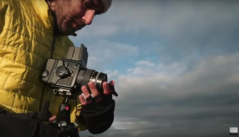 Photographer in yellow jacket holding a film camera outdoors under cloudy sky.