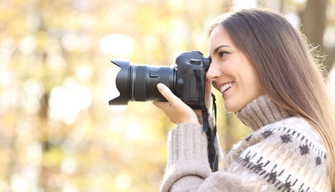Female photographer holding a DSLR camera to her face, smiling while composing a shot outdoors.
