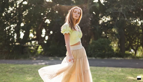 Woman in yellow crop top and beige tulle skirt posing outdoors in dappled sunlight.