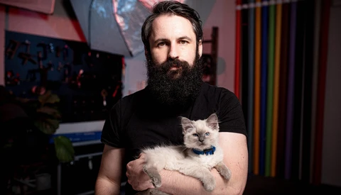 Bearded man in black t-shirt holding a light-colored kitten with blue collar in studio setting.