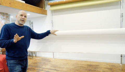 Man in navy sweatshirt gesturing toward blank whiteboard in bright studio space.