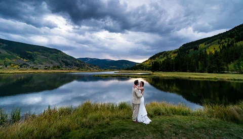 Couple in wedding attire standing in grass by a calm lake surrounded by forested mountains under dramatic cloudy sky.