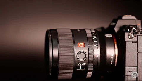 Sony mirrorless camera with attached telephoto lens featuring orange G branding against warm brown background.
