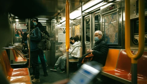 Passengers seated and standing inside a subway car with orange and green seating during daytime hours.
