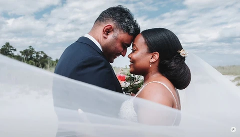 Bride and groom sharing an intimate moment outdoors, foreheads touching, with flowing veil in foreground.