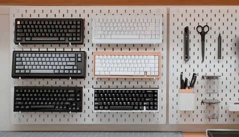 Pegboard wall display with six mechanical keyboards in various colors and a small tool organizer.