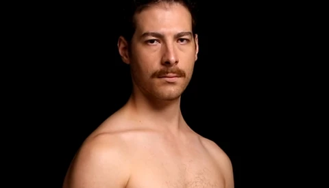 Shirtless man with dark hair and mustache against black background, looking toward camera.