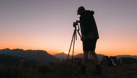 Silhouetted photographer on tripod against dramatic golden sunset sky with mountain range in background.