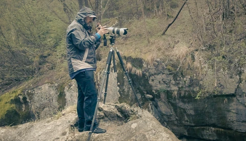 Photographer on rocky outcrop using telephoto lens mounted on tripod, woodland ravine in background.