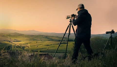 Photographer on a hillside at golden hour, aiming a telephoto lens on a tripod toward a scenic valley landscape.