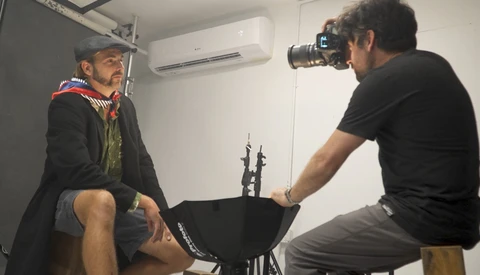 Photographer with camera on tripod conducting a portrait shoot with subject wearing cap and layered clothing.