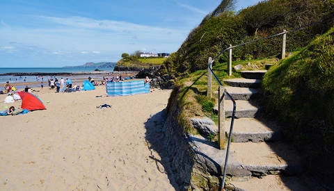 Wooden steps with rope railings descending a grassy cliff to a crowded beach with swimmers and colorful umbrellas.