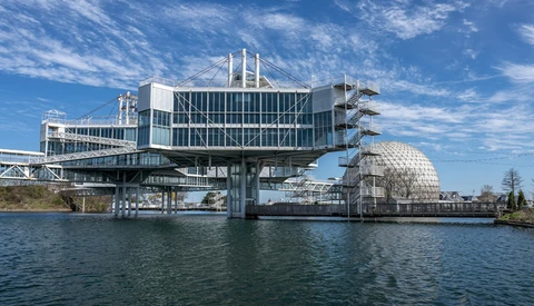 Modern waterfront building with white vertical columns and glass facade cantilevers over calm blue water.