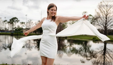 Woman in white dress holding flowing white fabric near a calm lake with bare trees reflected in the water.