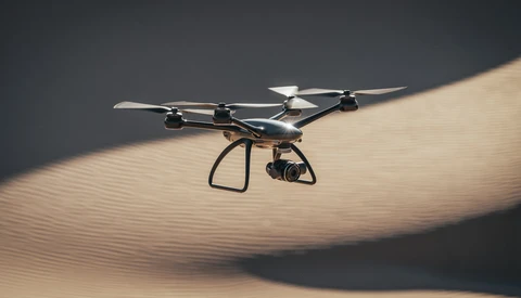 Quadcopter drone hovering above rippled sand dunes with dramatic side lighting casting sharp shadows.