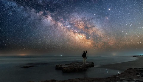 Silhouetted figure standing on a rocky outcrop facing a vast starry night sky with the luminous Milky Way galaxy arching overhead.