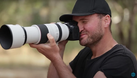 Photographer holding a telephoto lens with white barrel, looking through viewfinder in outdoor setting.