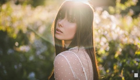 Young woman with long dark hair and bangs looking over her shoulder in a sunlit wildflower field.