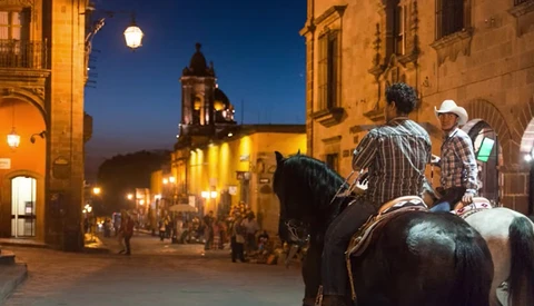Two horseback riders in cowboy hats on a colonial town plaza at dusk, with illuminated historic buildings and a church tower in the background.