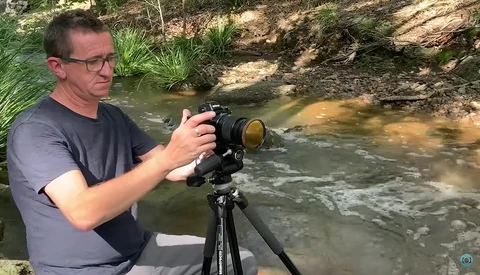 Photographer with telephoto lens on tripod aiming toward a flowing stream in a wooded area.