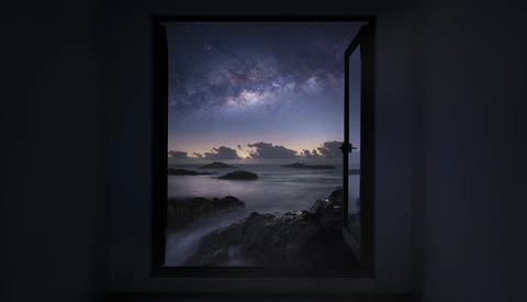 Open window framing a coastal night scene with the Milky Way visible in the starry sky above a calm sea.