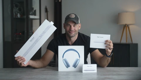 Man in black shirt and cap displays unboxed wireless headphones and packaging materials at a table.