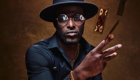 Man wearing black hat and round glasses gestures expressively against warm brown backdrop.